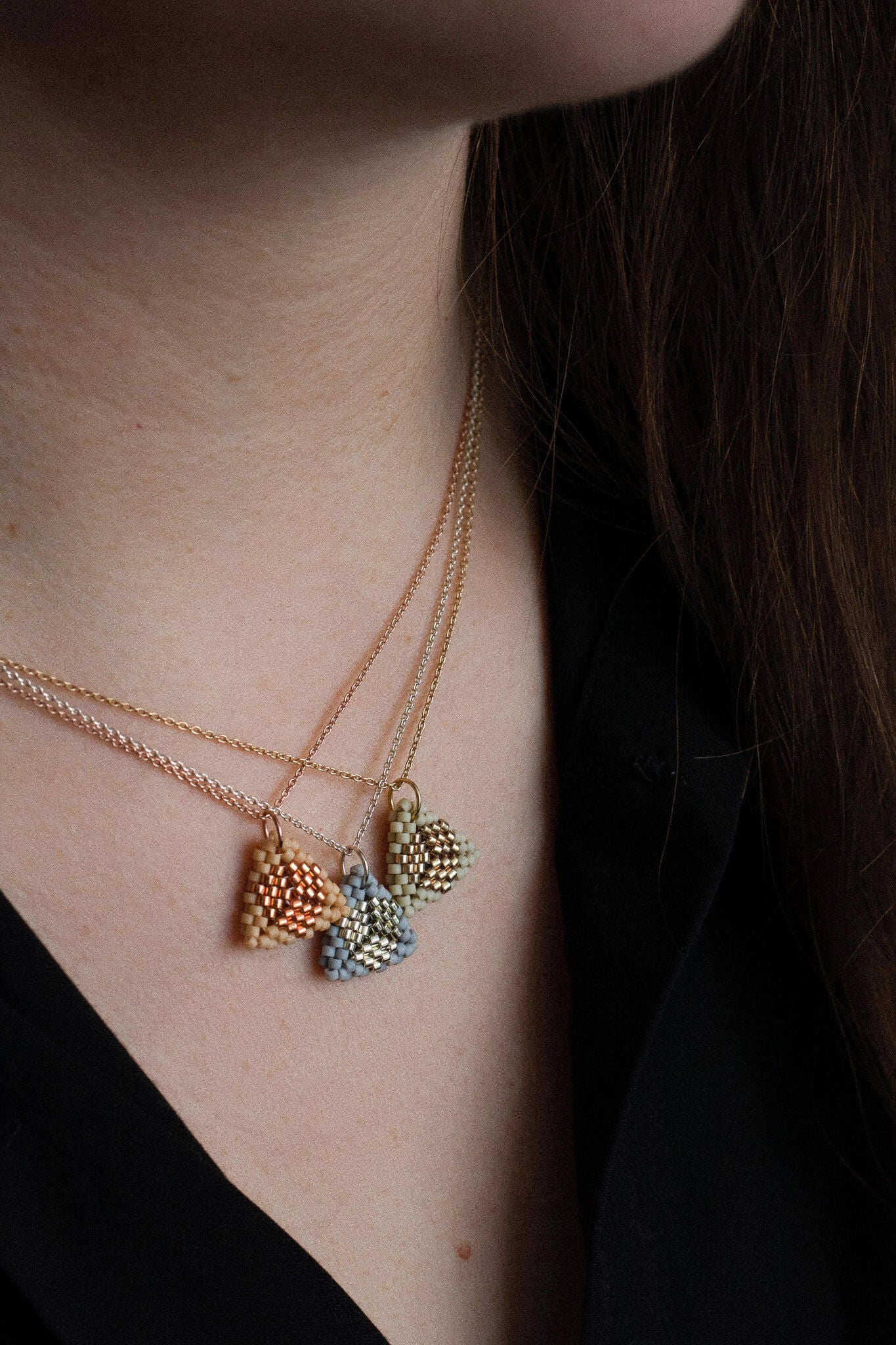 Three beaded triangle pendant necklaces in beige, gray and breige tones, on silver, gold and rose gold chains, worn layered on neck.