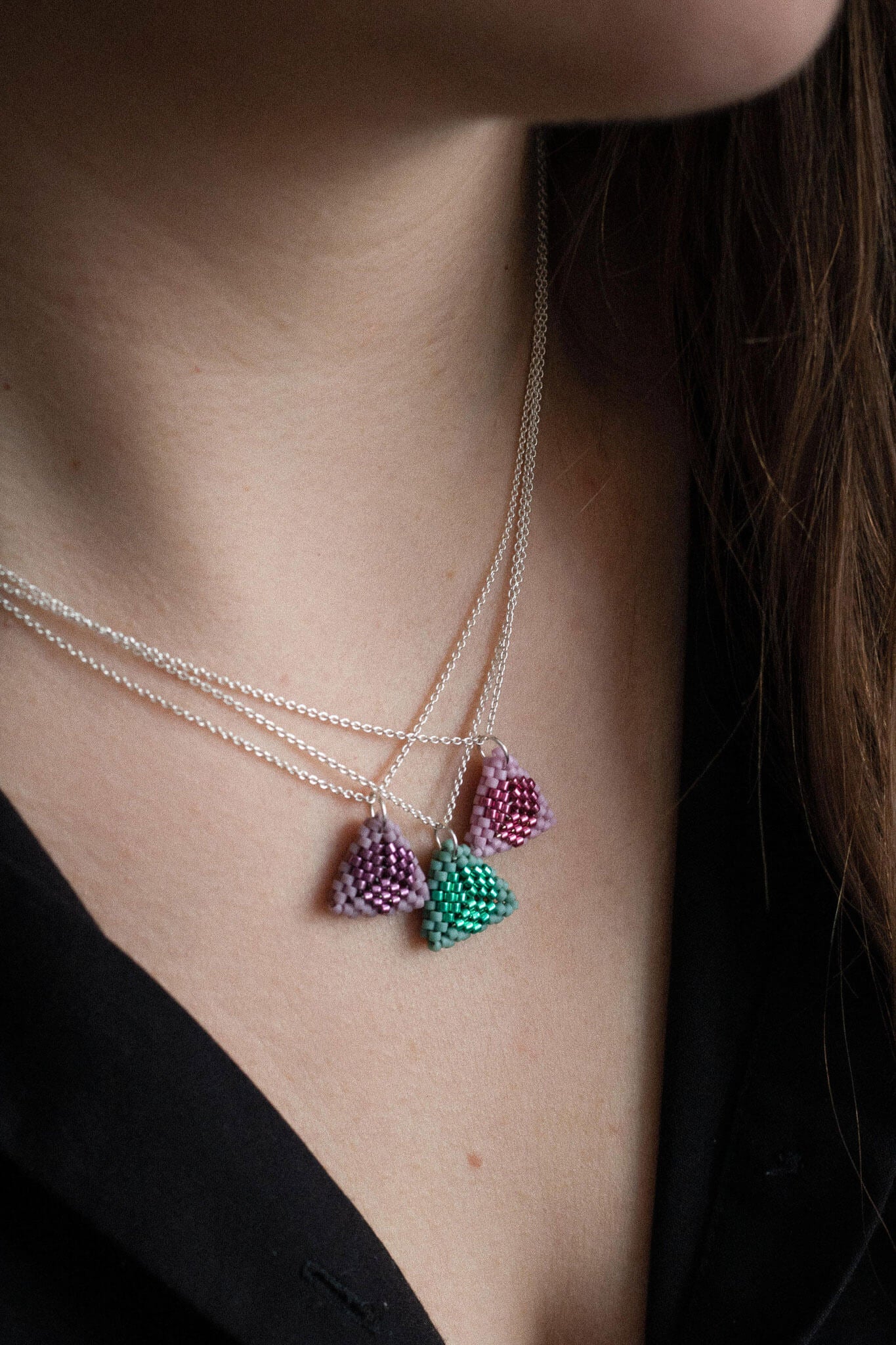 Three beaded triangle pendant necklaces in pink, purple and green tones, on silver chains, worn layered on neck.