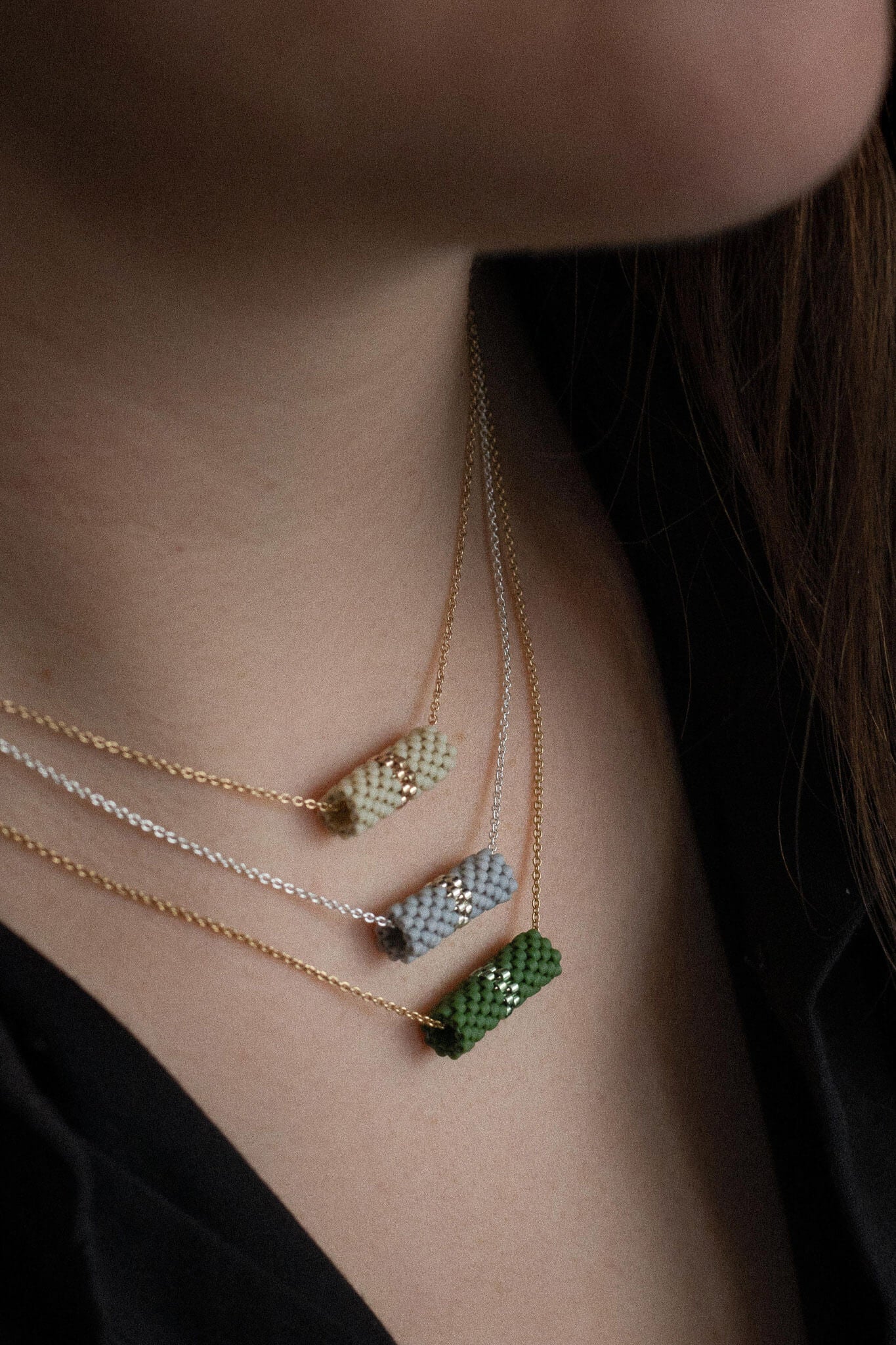 Three beaded tube pendant necklaces in green, gray, and beige tones on gold and silver chains worn layered on neck.